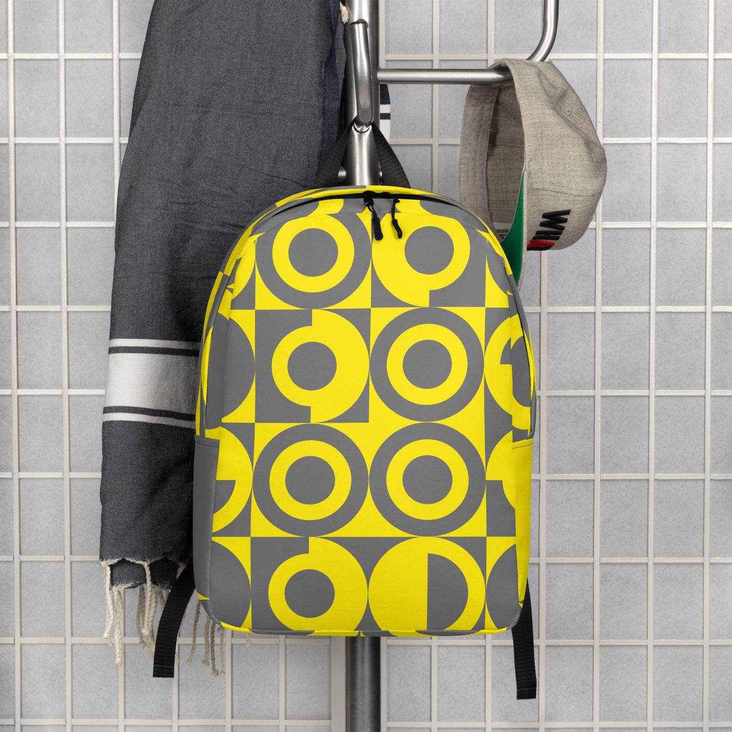Yellow and Grey Geometric Pattern Backpack, front view with a grey tile background hanging on a silver coat rack with a grey hat and blanket