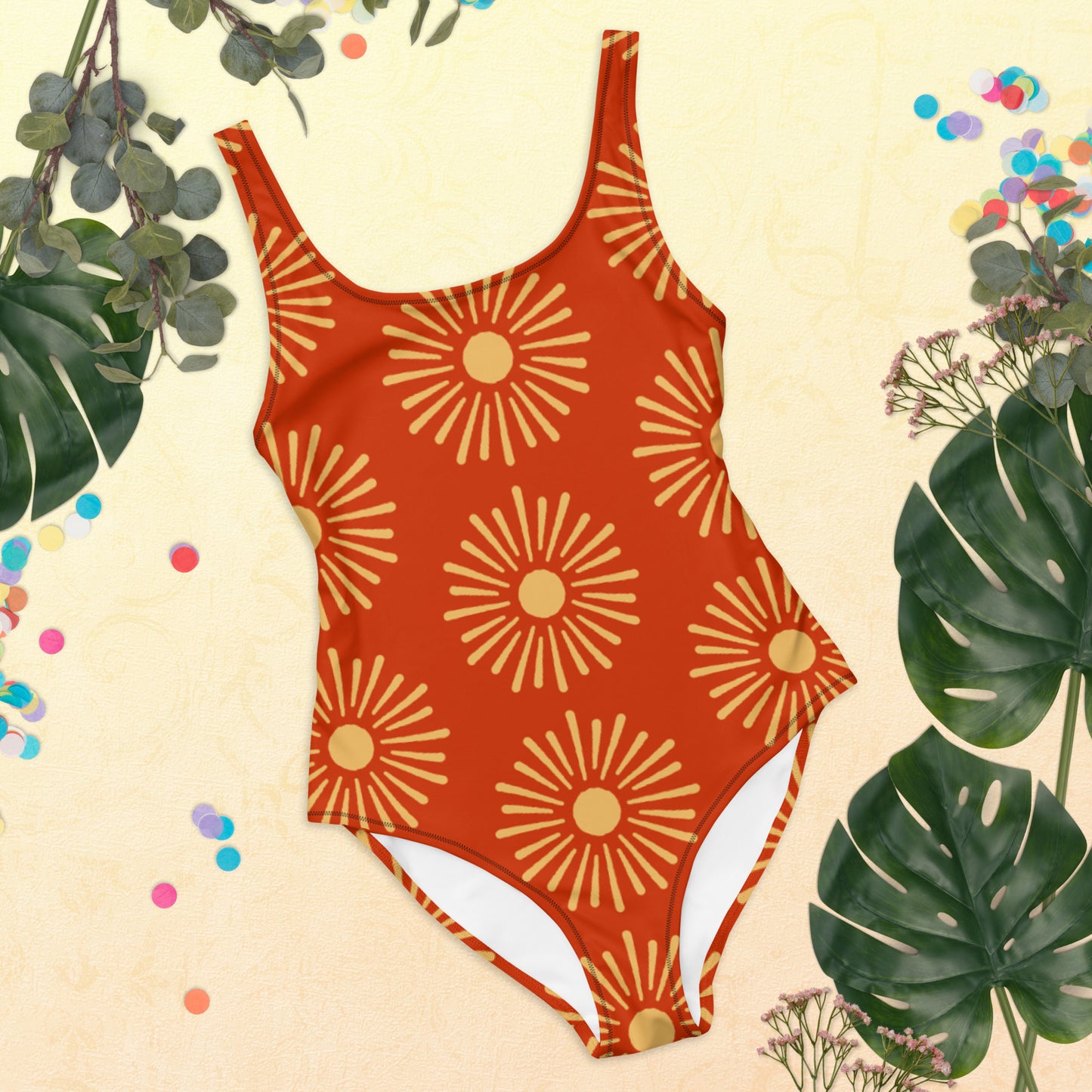 all-over-print-one-piece-swimsuit in Terra-cotta with Yellow sun Print, By itself laying flat on a table with plant leaves surrounding it