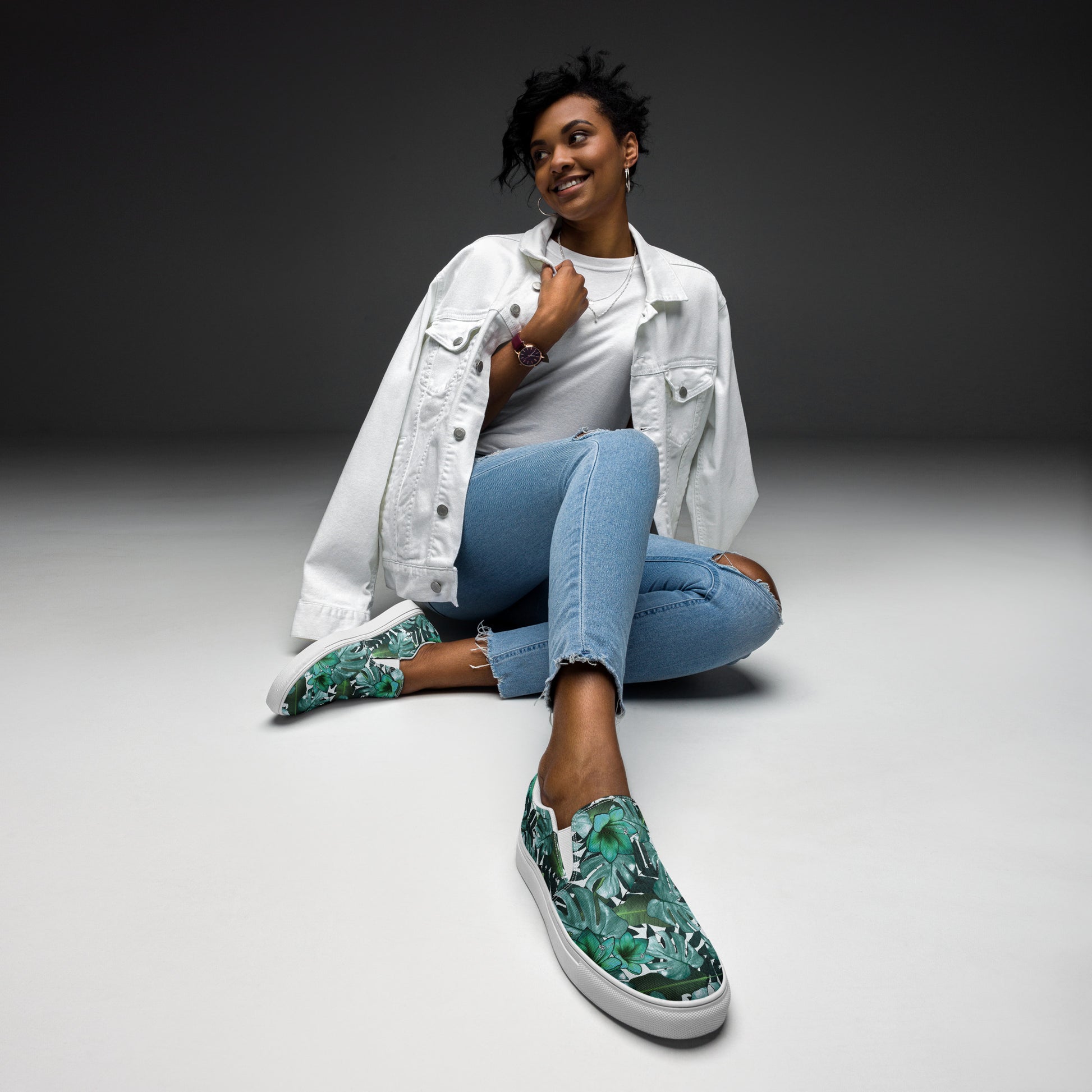multicolor green tropical print canvas slip on shoes top view while a woman is sitting on the ground dresses in jeans and a white jacket