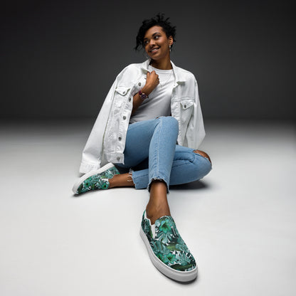 multicolor green tropical print canvas slip on shoes top view while a woman is sitting on the ground dresses in jeans and a white jacket