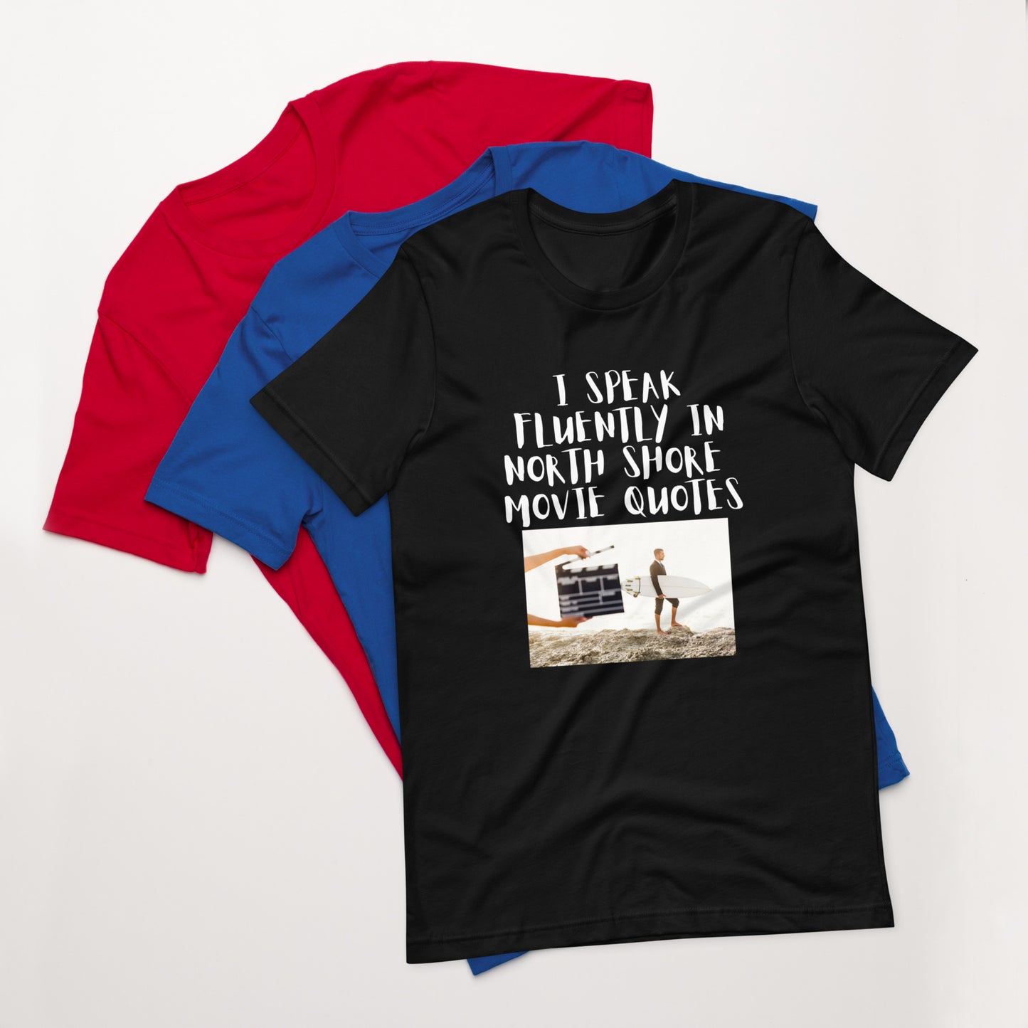 I speak in North Shore Movie Quotes in large text, Picture of a Movie Scene Marker with Surfer holding a surfboard on the beach short sleeve t-shirt in black, blue and red laying flat 