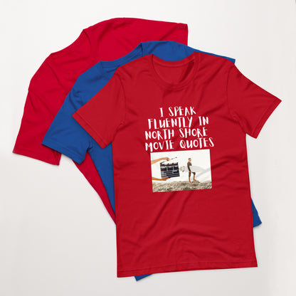 I speak in North Shore Movie Quotes in large text, Picture of a Movie Scene Marker with Surfer holding a surfboard on the beach short sleeve t-shirt in Red, laying flat ontop of a red and blure shirt